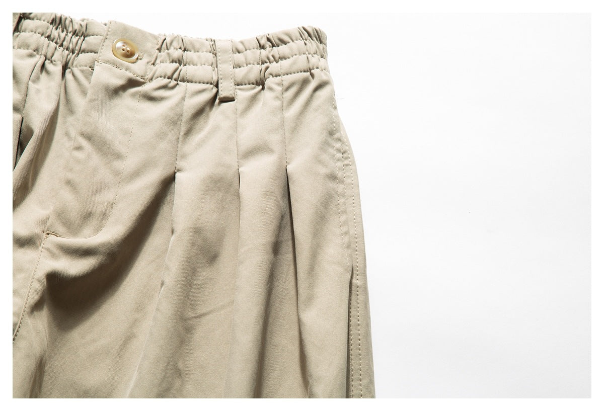 Close-up of light beige pants showing the elasticated waistband, button, belt loop, pleats, and side seam.