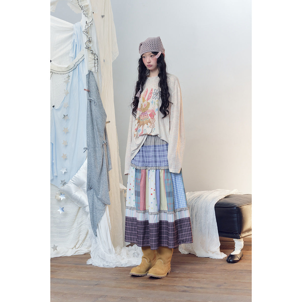 Model wearing a cream illustrated long-sleeve top, a tiered blue and multicolor patchwork skirt, a mauve knit cap and tan