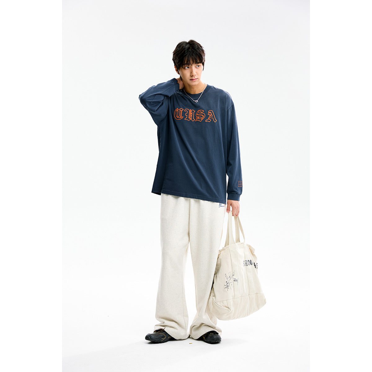Model wearing a navy long-sleeve shirt with orange USA lettering and cream wide-leg pants, holding a canvas tote.