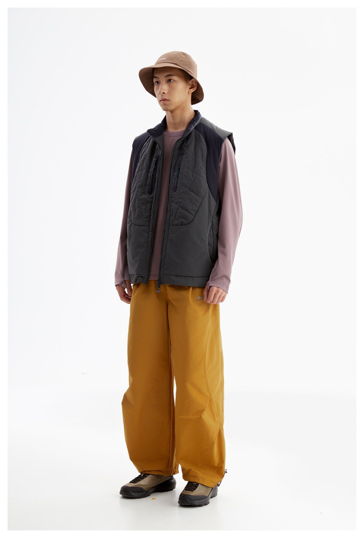 Model wearing a charcoal quilted utility vest over a mauve shirt, mustard wide-leg pants, and a tan bucket hat.
