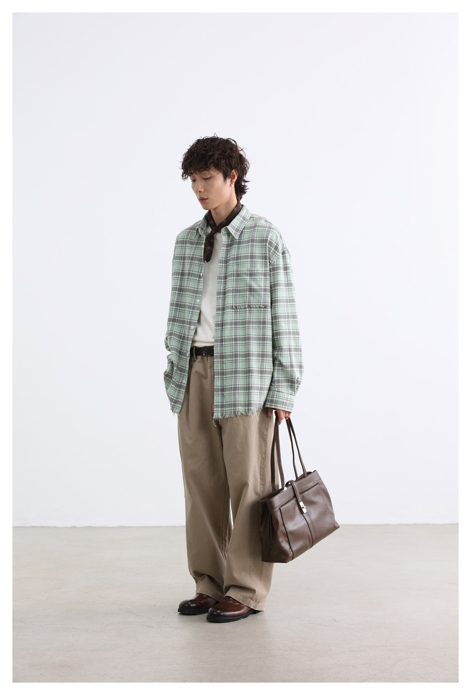 Model wearing a green plaid shirt open over a white top with khaki pants and holding a brown tote bag