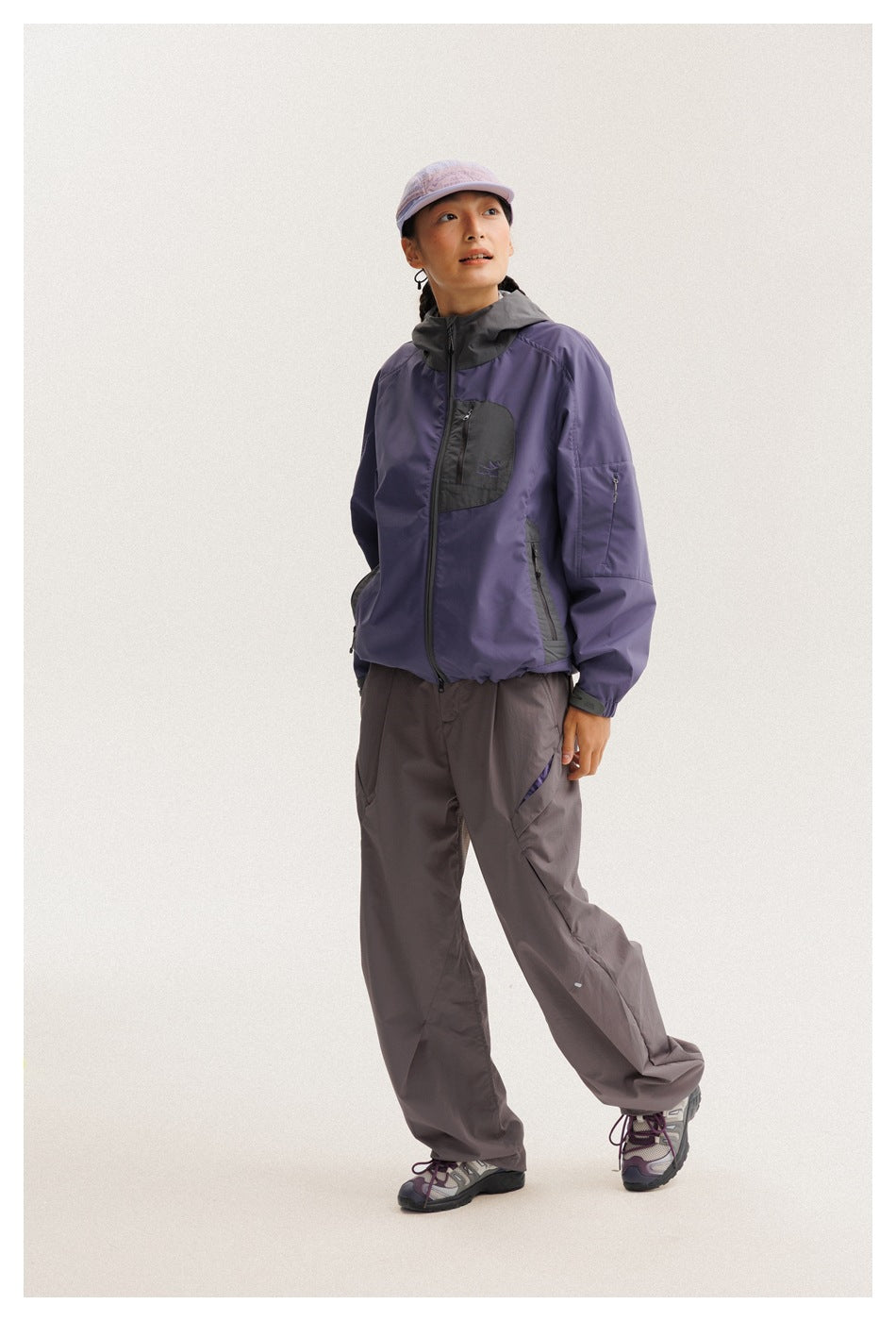 Full-length view of a model wearing a purple and charcoal hooded jacket, loose gray pants, and a lavender cap.