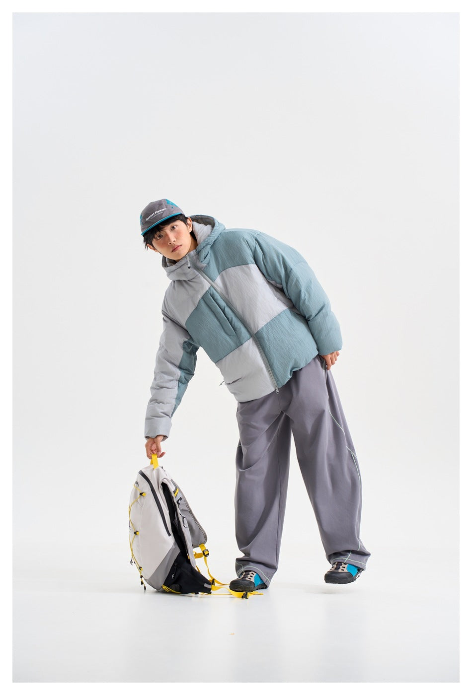 A model leans beside a backpack, wearing a pale blue, gray, and white color-block hooded puffer jacket.