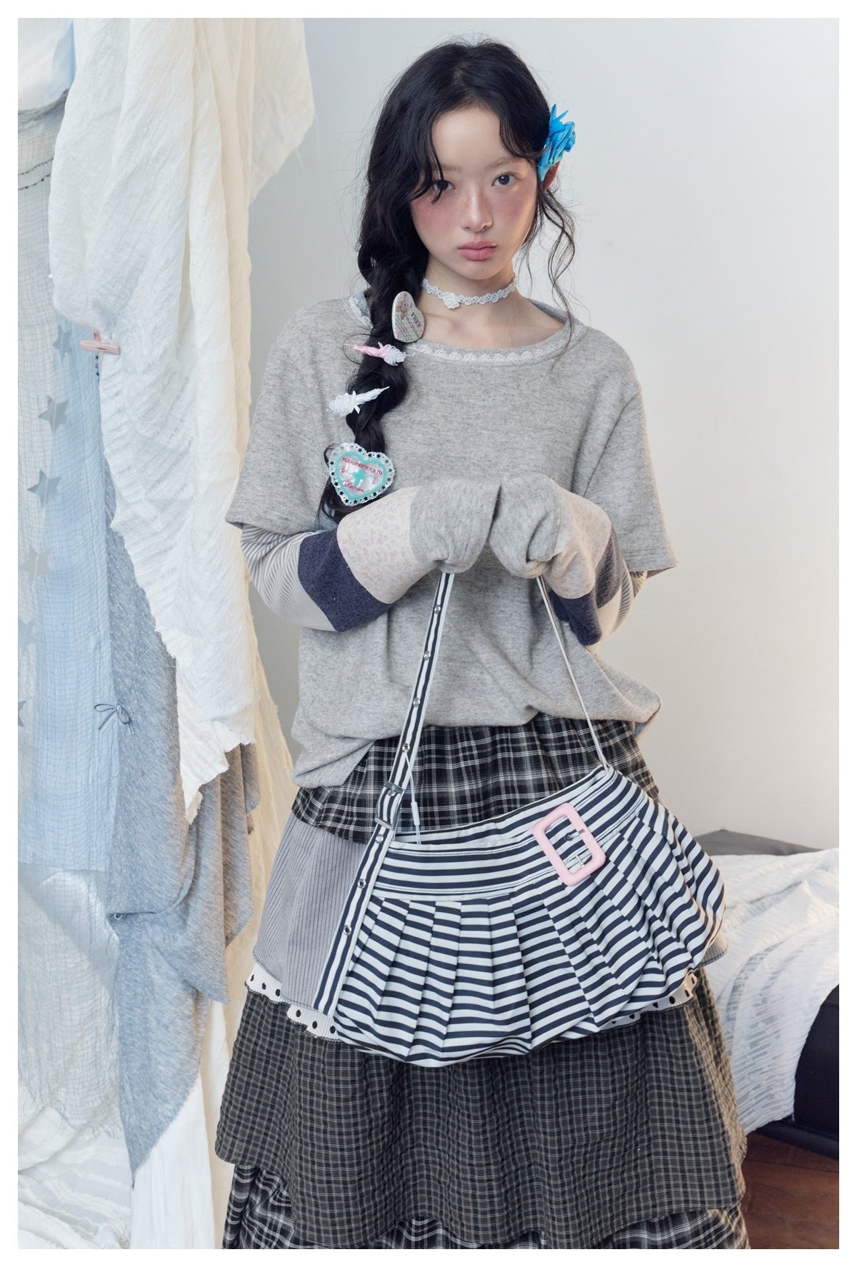 Model wearing a loose gray lace-trim top with layered sleeves, a dark plaid skirt, and a striped shoulder bag.