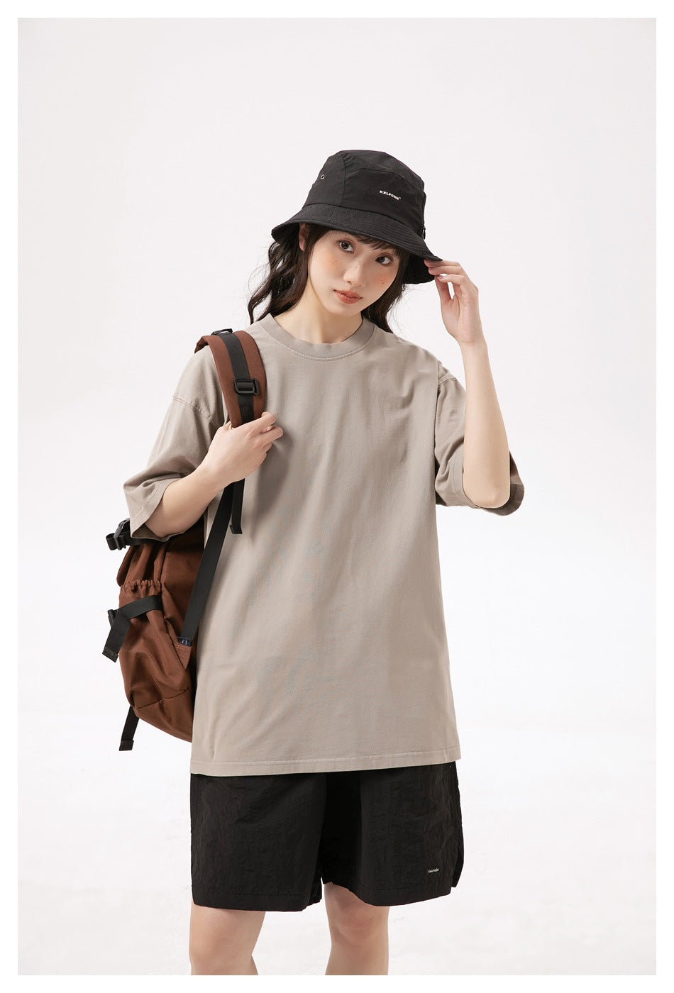 Model wearing an oversized taupe T-shirt with black shorts, a black bucket hat, and a brown backpack.
