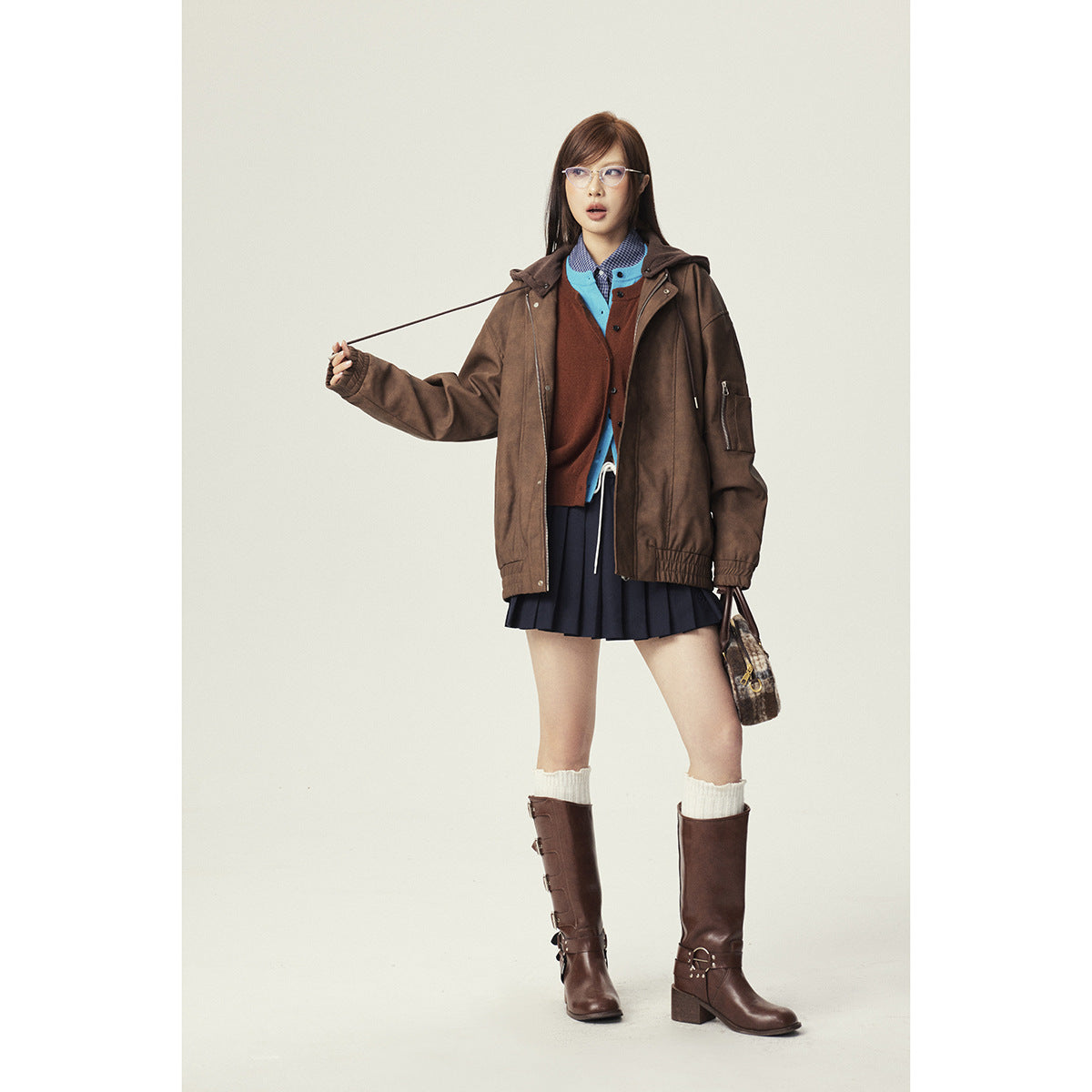 Model wearing an oversized brown hooded jacket over layered tops and a navy pleated skirt, with brown boots.