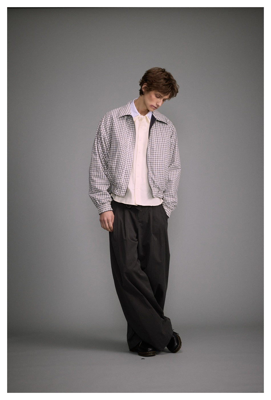 Full-length model wearing a light plaid zip-front jacket over a pale shirt and loose black trousers.