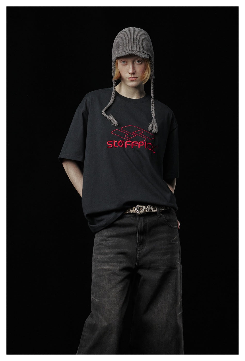 Model wearing a black T-shirt with red chest lettering, dark wide-leg jeans, and a gray knit cap with braided ties.