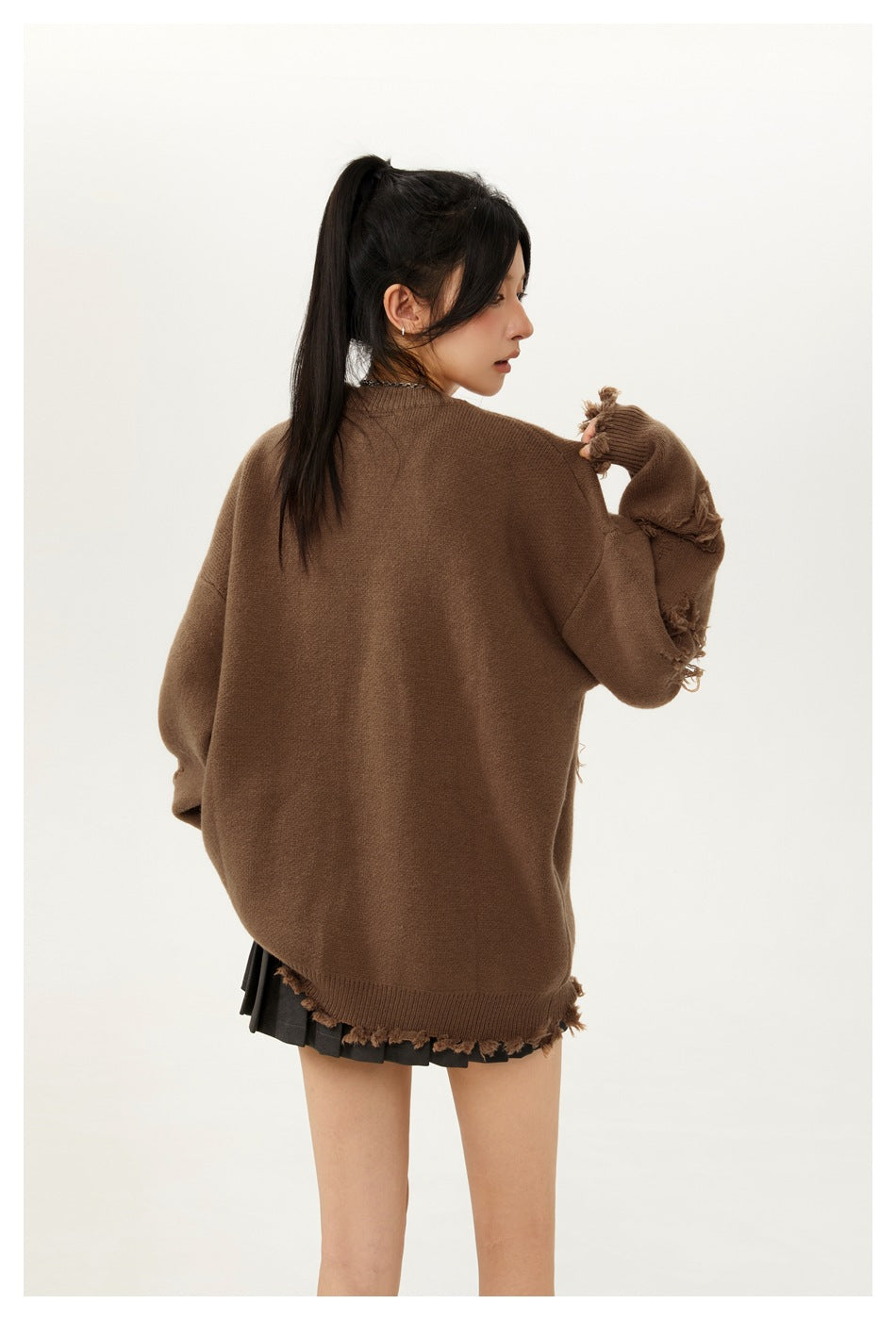 Model showing the back of an oversized brown sweater with distressed sleeve details and a black pleated mini skirt.