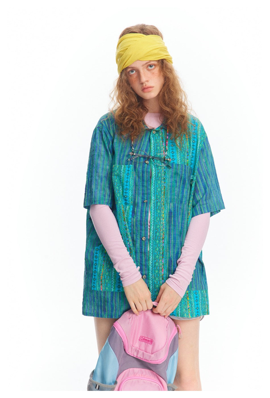 Model wearing a blue-green patterned short-sleeve button shirt over pink sleeves, with a yellow headband.
