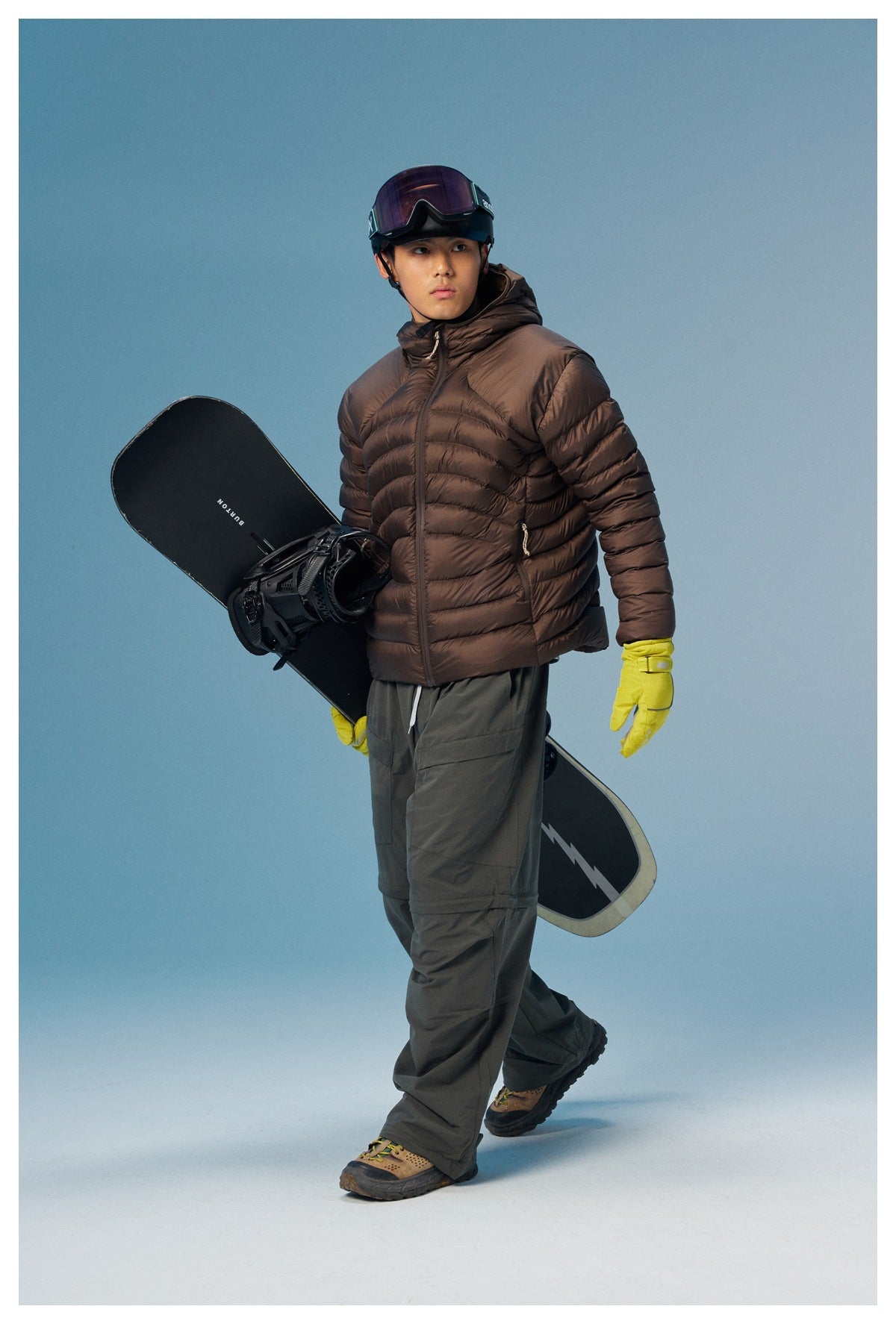 Full-length model wearing a dark brown quilted hooded jacket, holding a snowboard.