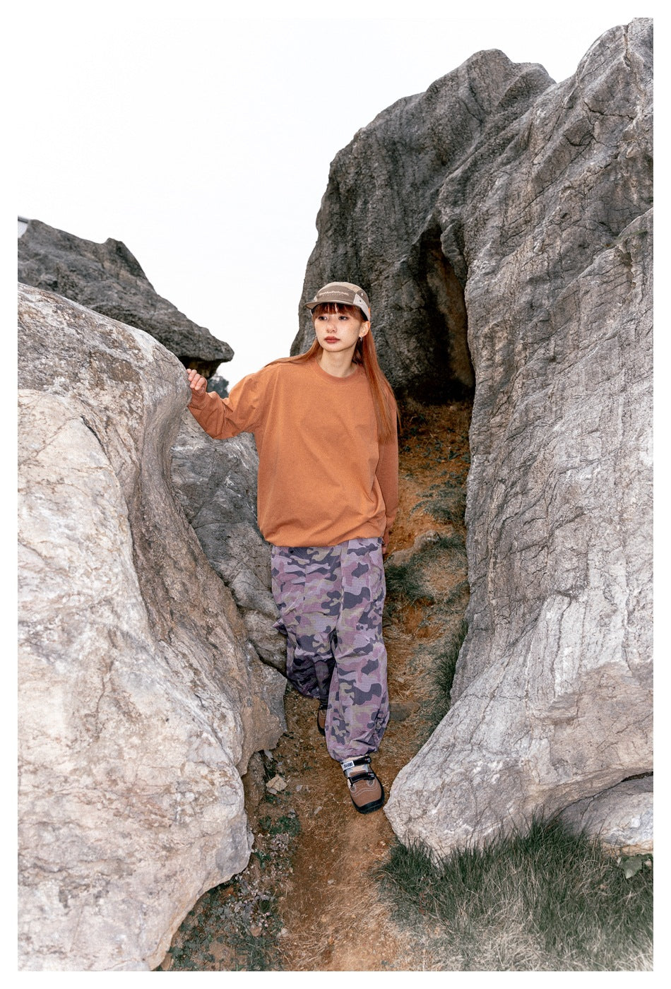 Model in a rust-orange sweatshirt and purple camouflage pants walking between large rocks.