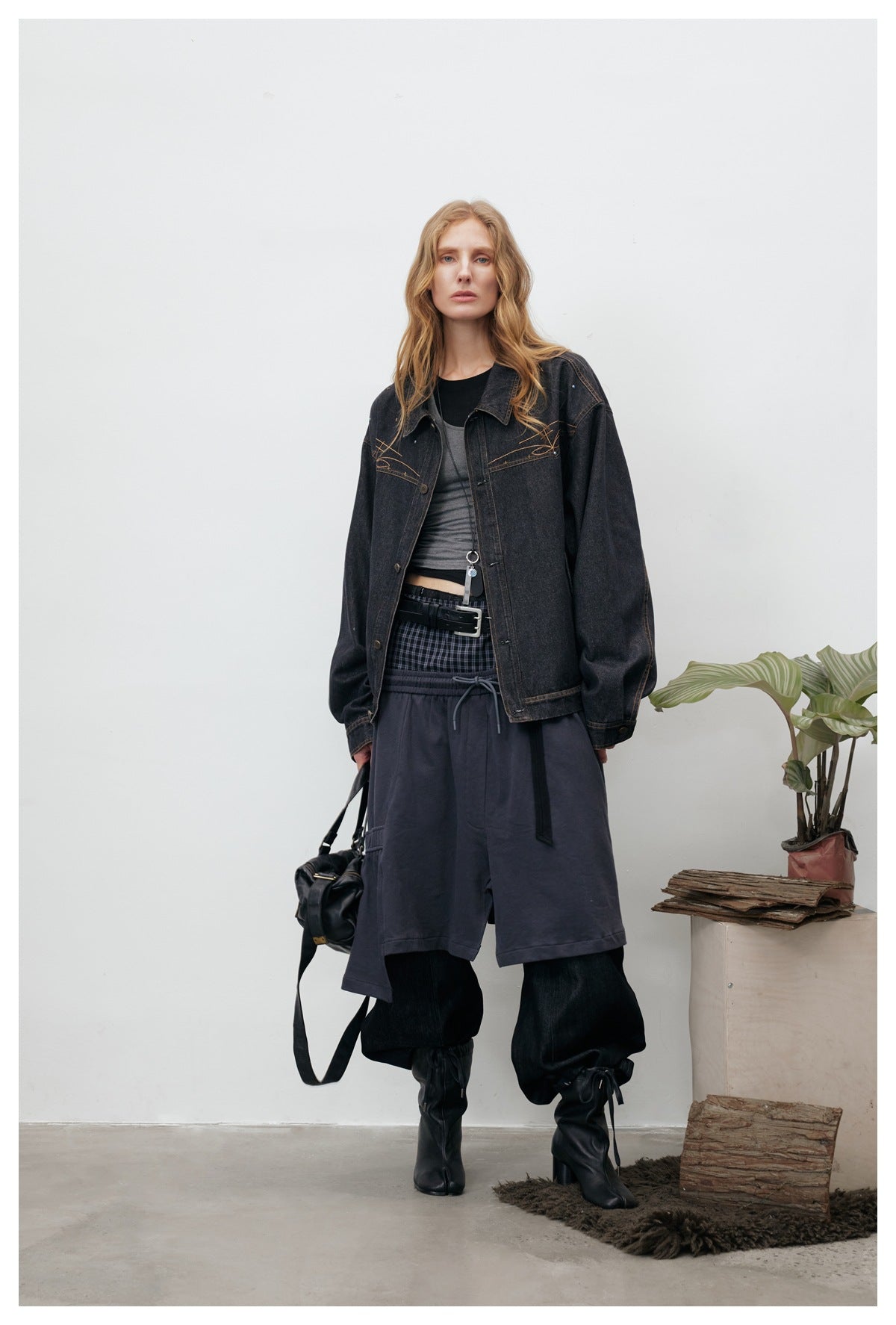 Model wearing a washed black embroidered denim jacket open over a gray top, with a layered dark skirt, black boots and a bag