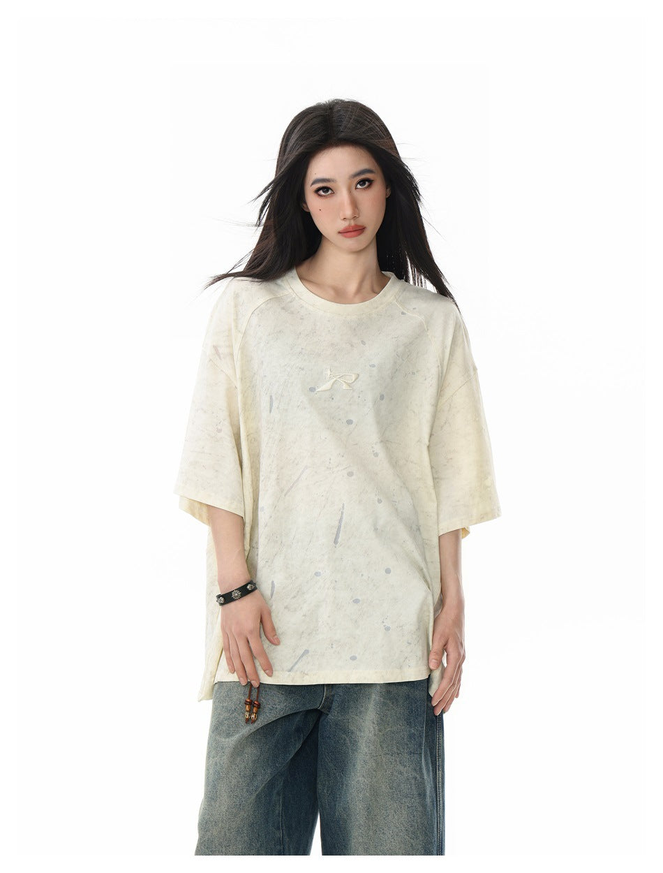 Model wearing oversized beige crew-neck T-shirt with gray splatter marks and small logo, styled with faded blue jeans