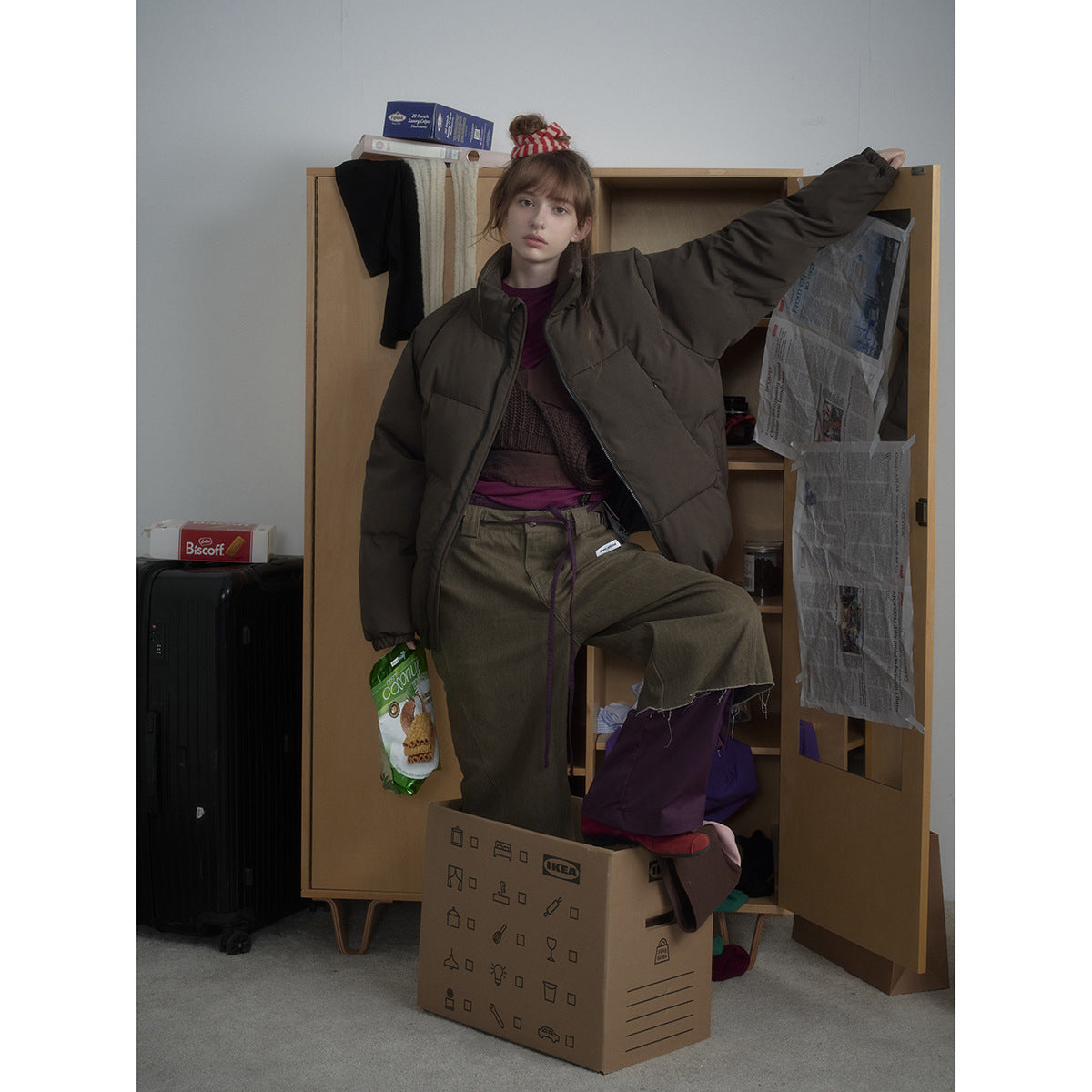 Model wearing an oversized dark olive-brown puffer jacket with wide olive trousers, posed in front of an open wardrobe.