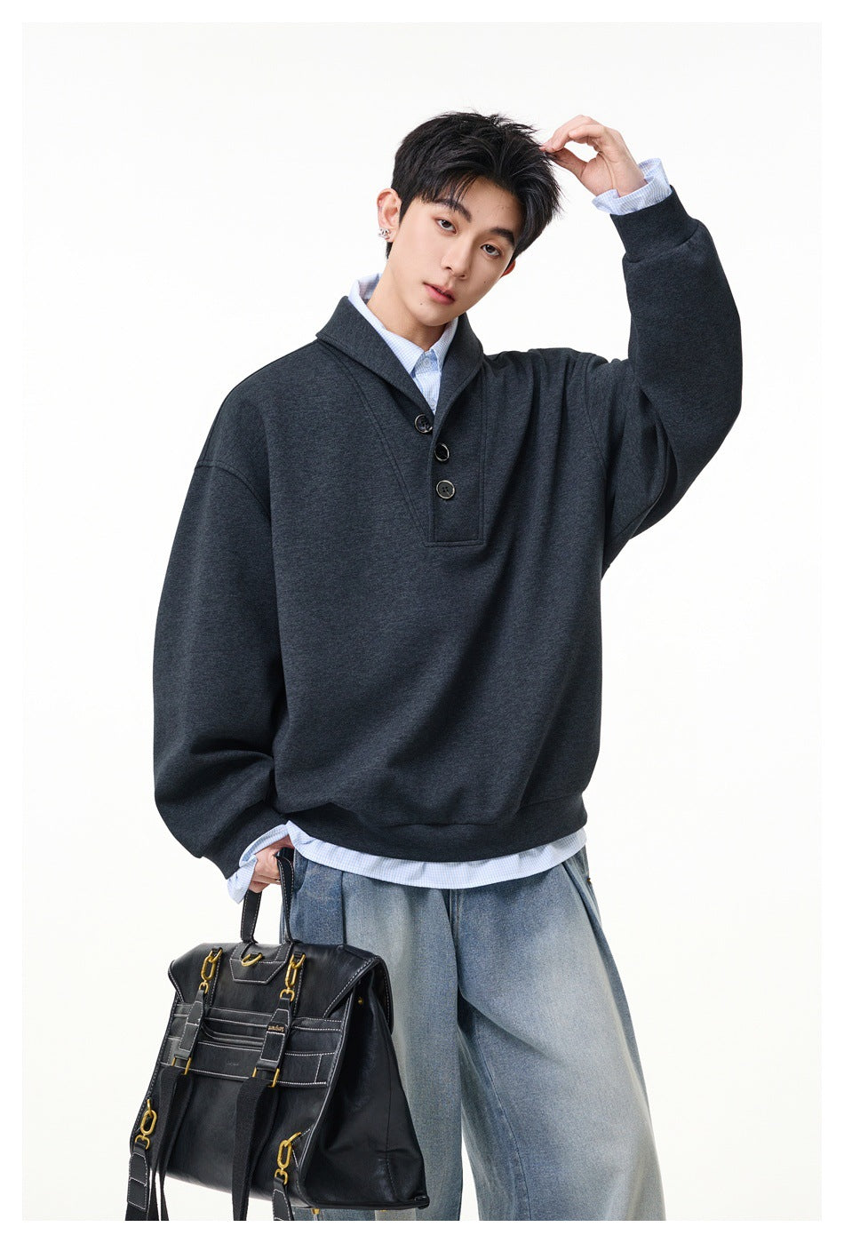 Man wearing a dark gray shawl-collar sweatshirt with buttons over a blue gingham shirt, with wide jeans and a black bag