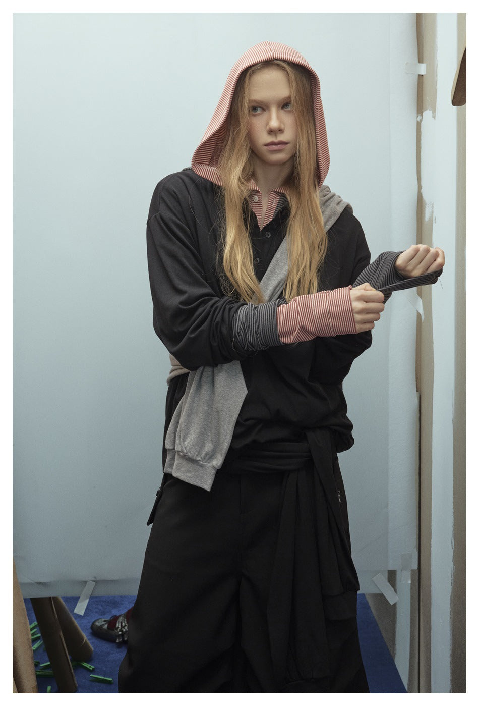Model in a black hooded top with red striped hood and cuffs, gray layer tied across the body, and black bottoms.