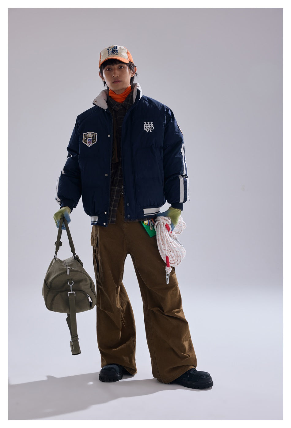Model wearing navy puffer jacket with chest patch and sleeve stripes, brown cargo pants, orange cap, holding a duffel bag