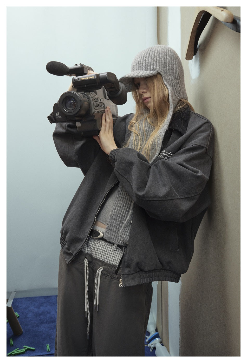 Model in a charcoal oversized jacket and dark pants, wearing a gray knit cap and scarf while using a video camera.