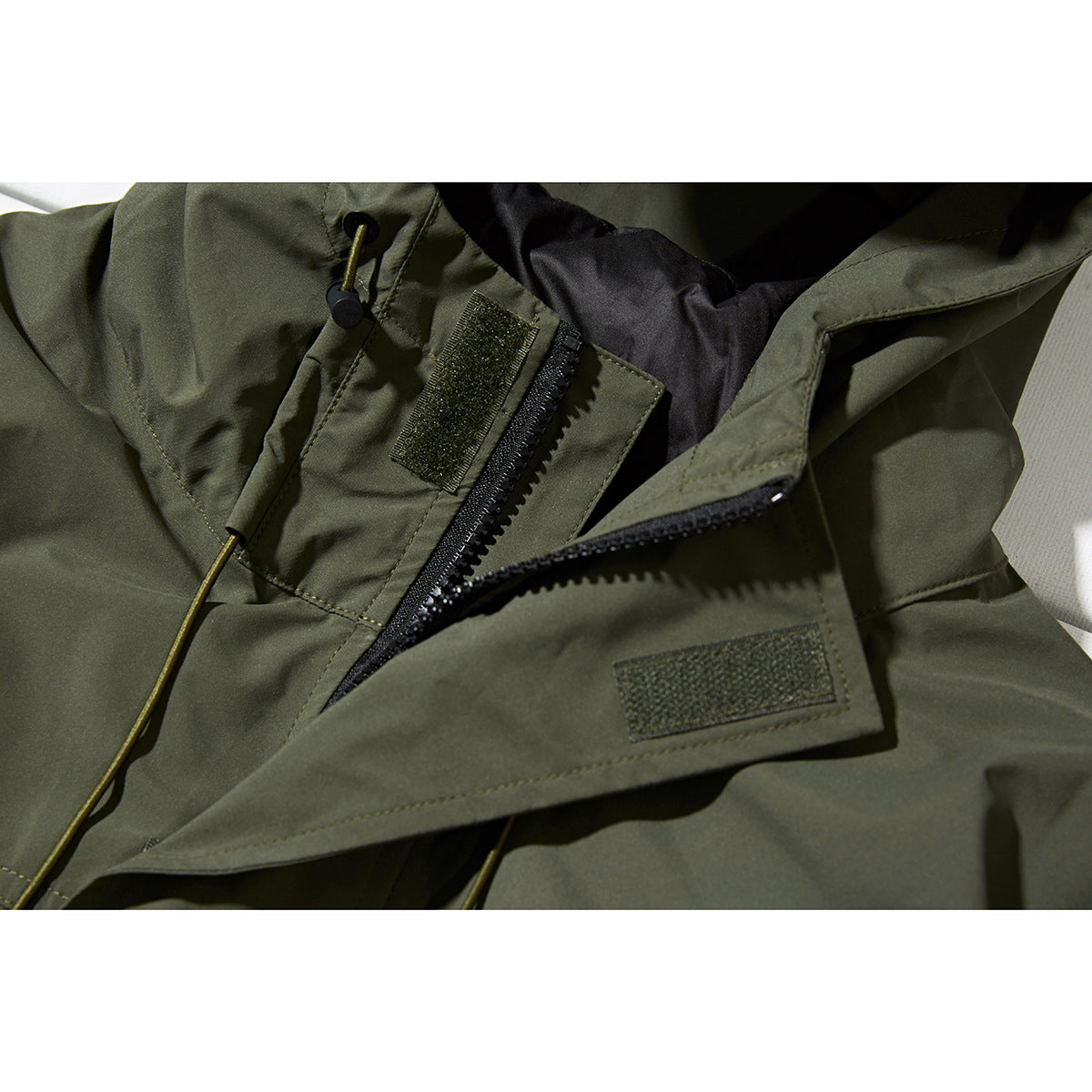 Close-up of an olive jacket's front zipper, hook-and-loop storm flap, and hood drawcord.