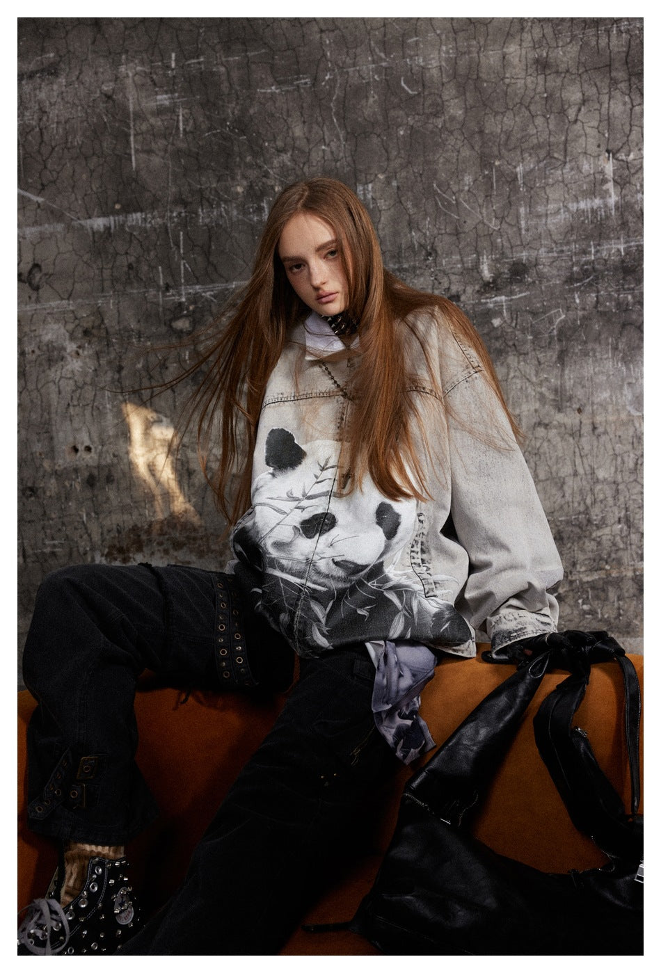 Model seated wearing loose gray panda-print jacket with black pants against a cracked concrete wall