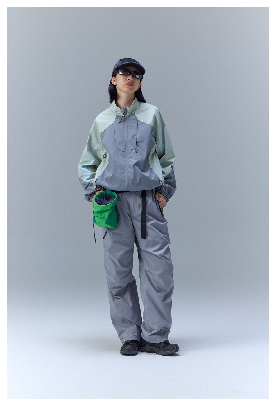 Model wearing a pale mint and blue-gray color-block zip jacket with gray pants and a dark cap.