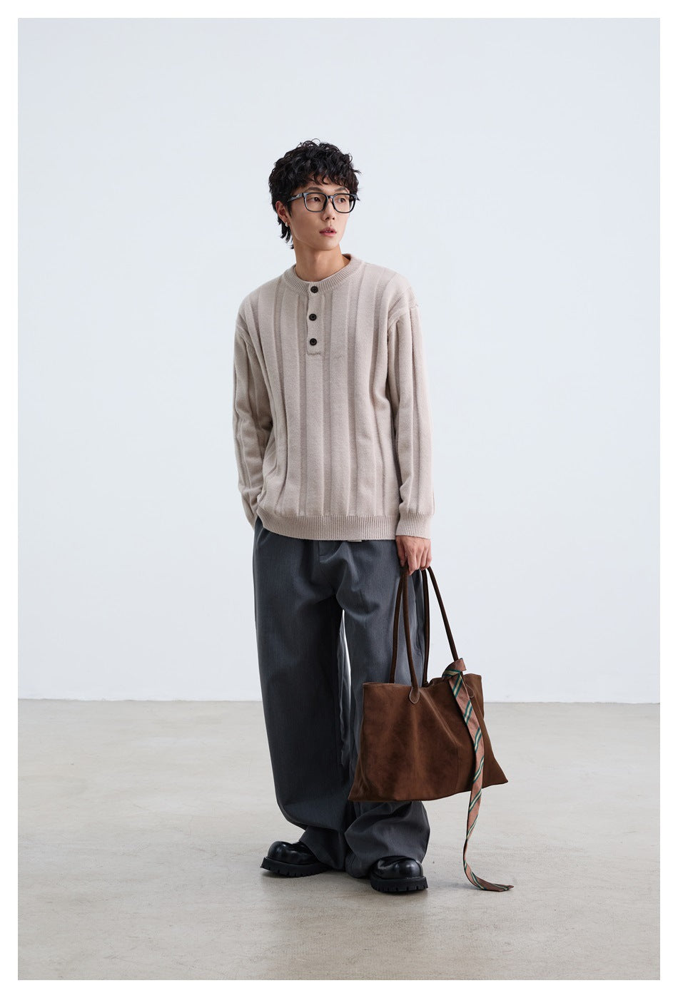 Full-length model wearing a pale beige ribbed button-neck sweater with dark trousers and carrying a brown tote.