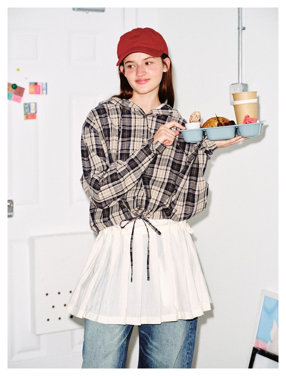 Model wearing a gray plaid hooded button-up with a drawstring hem and white pleated layer, shown from the waist up.