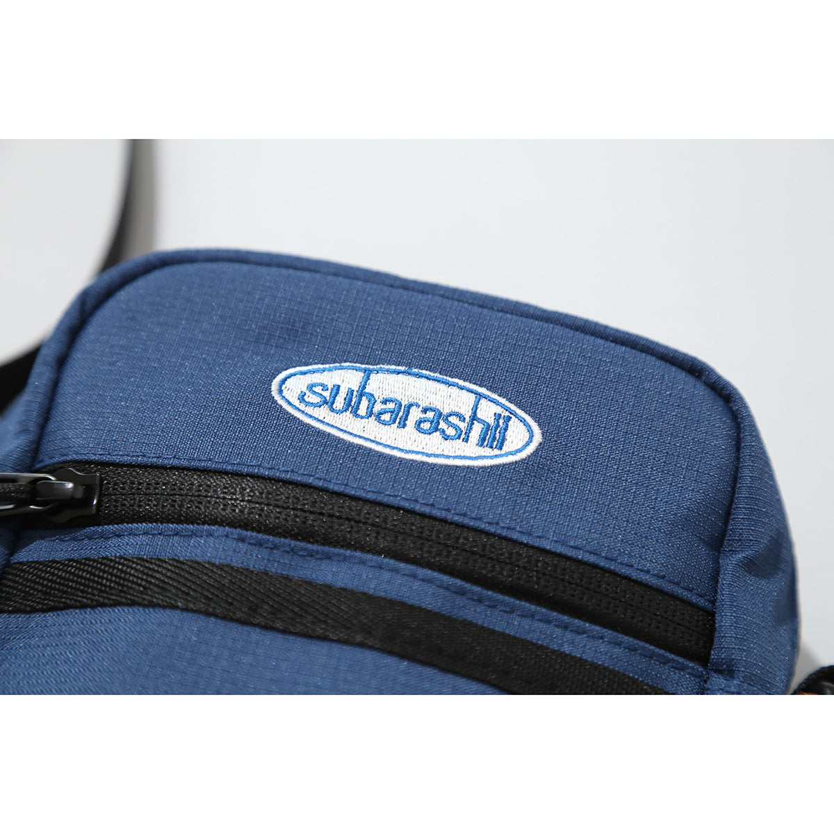 Close-up of the navy bag's embroidered oval logo, black zipper, and textured fabric.