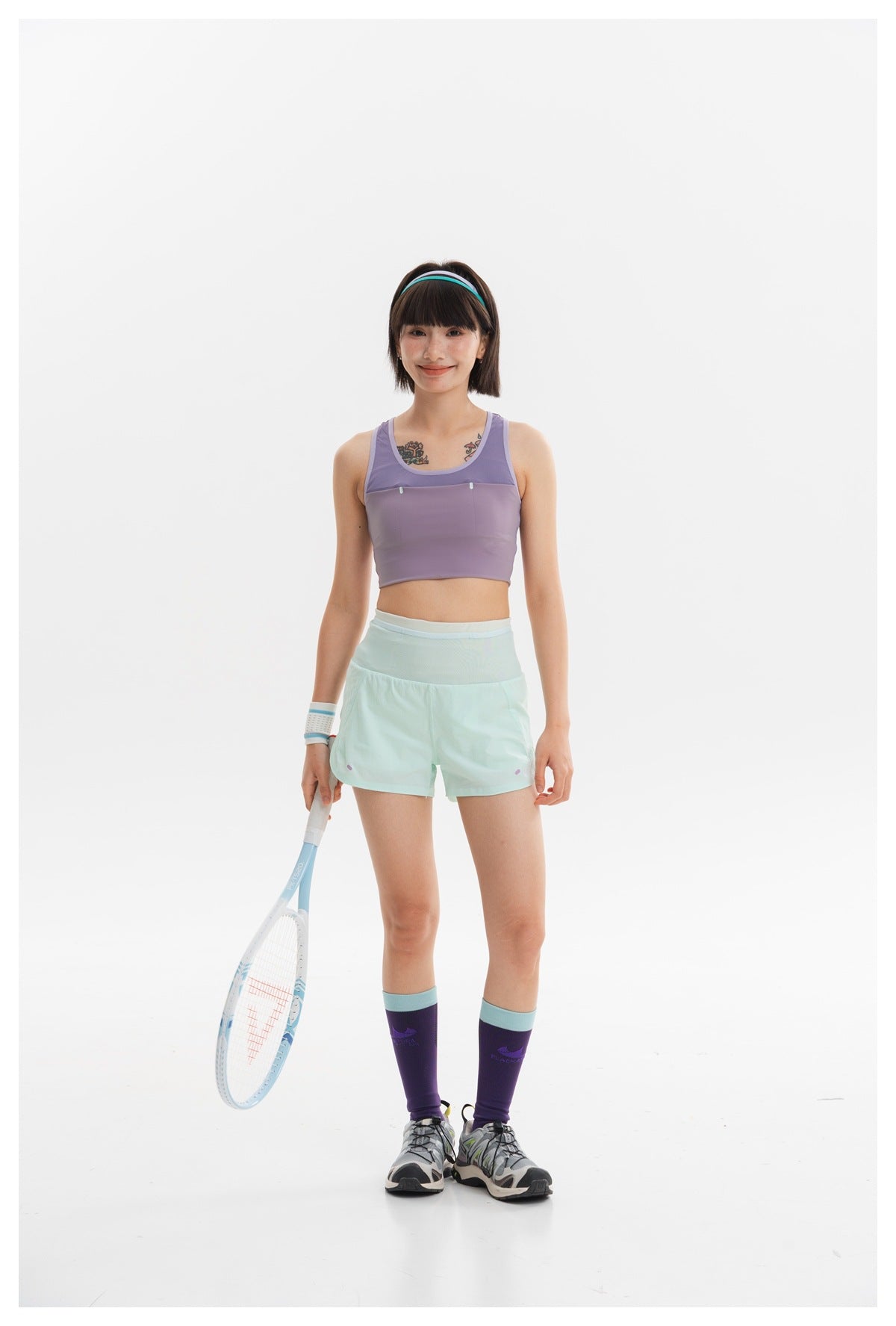 Model wearing a lavender cropped sports top and mint shorts, holding a tennis racket against a white background.