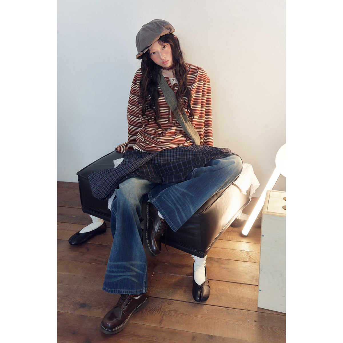 Model seated in a rust, brown and cream striped long-sleeve henley with wide-leg jeans and a plaid garment across her lap.