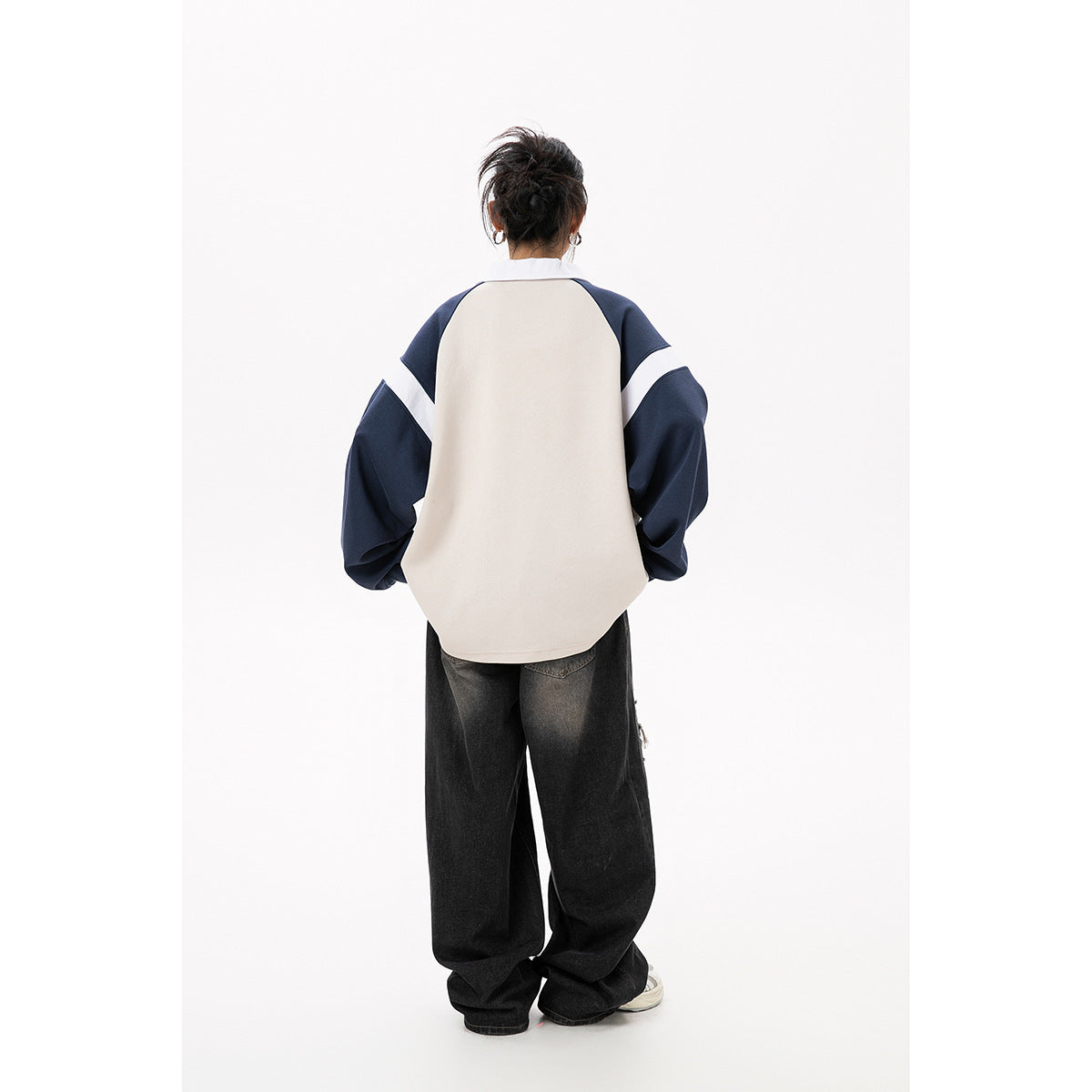Back view of a model wearing a cream polo sweatshirt with navy sleeves and white arm stripes, paired with loose black jeans.