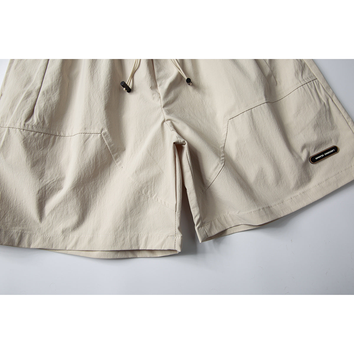 Close-up of cream shorts showing angled front panels, hem stitching, drawstrings, and a small dark leg patch.