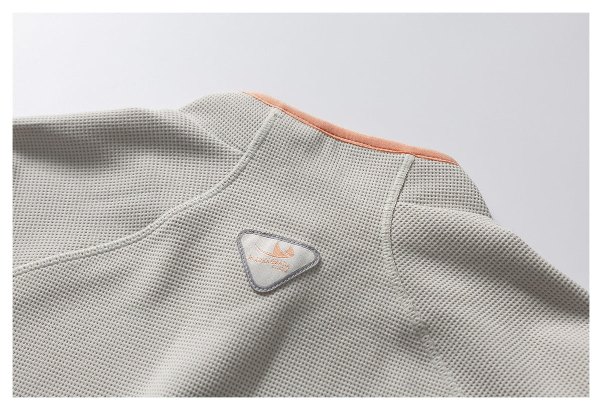 Close-up of a pale gray waffle-knit garment showing an orange-trimmed neckline and a small triangular stitched chest patch.