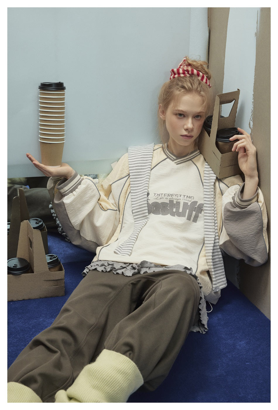 Model wearing a cream graphic pullover with gray piping and striped trim, loose dark trousers, and pale leg warmers.