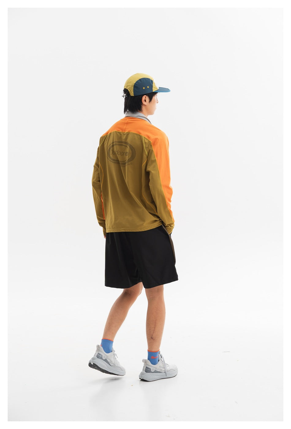 Rear three-quarter view of a model wearing an olive and orange color-block long-sleeve top with black shorts.