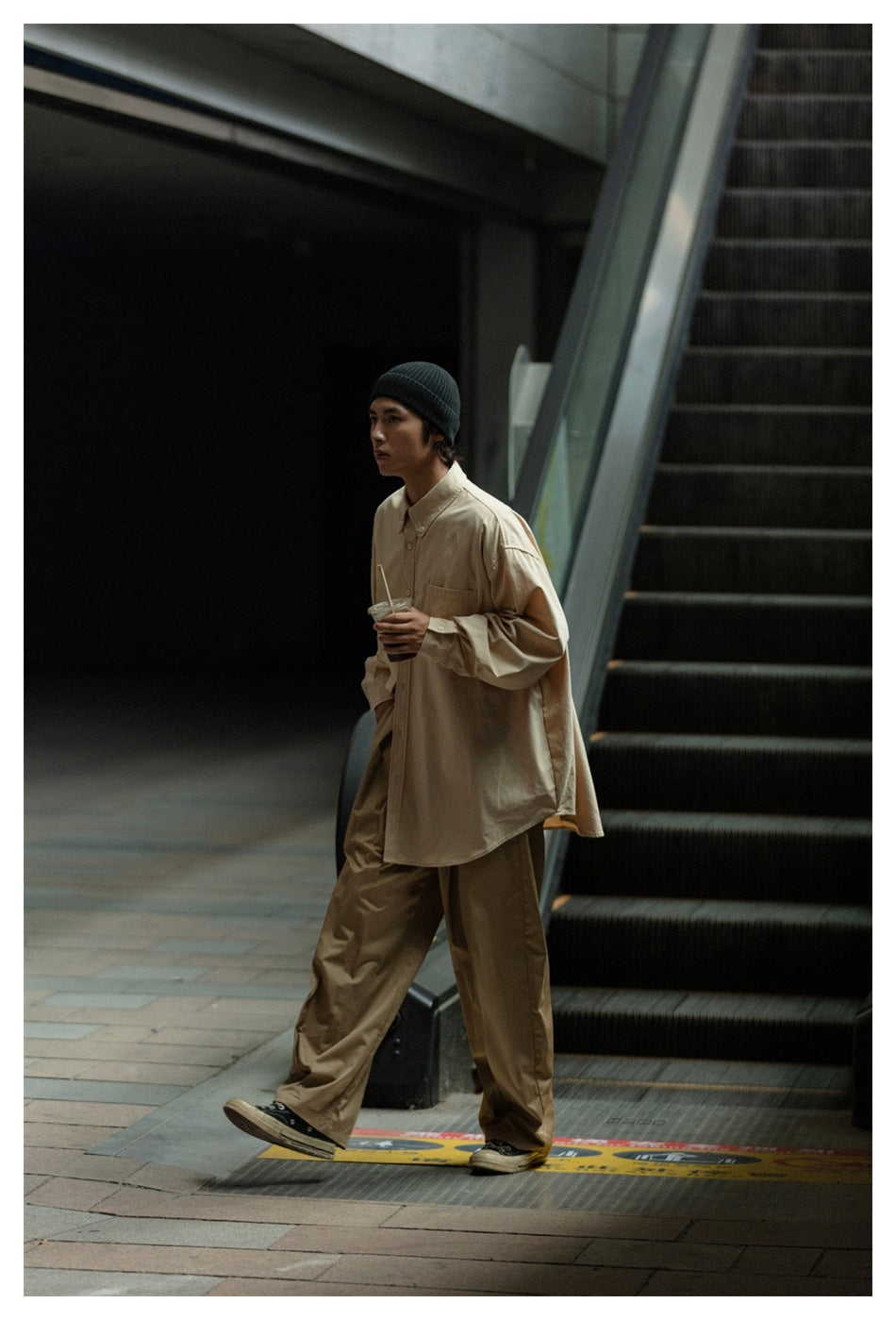 Model in a loose beige button-up shirt and wide tan trousers, walking beside an escalator.