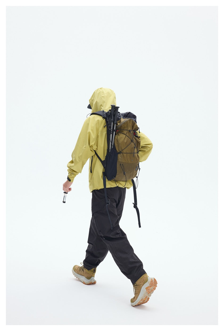 Rear view of a hiker in a yellow hooded jacket and black pants, carrying a brown backpack and trekking poles.