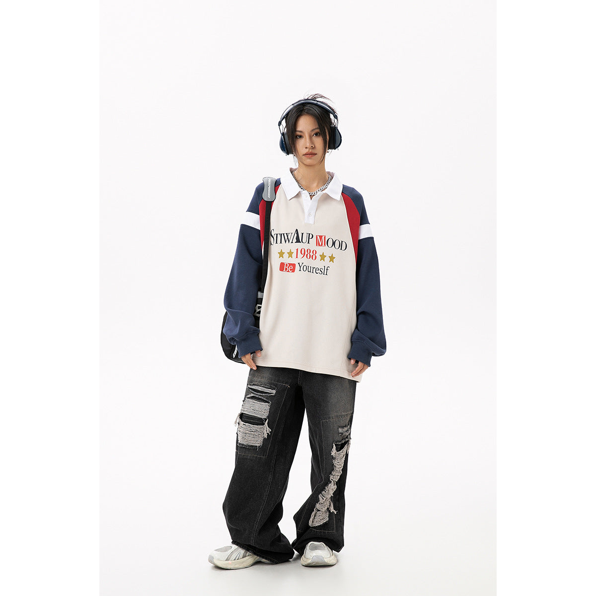 Full-length front view of a model wearing a cream polo sweatshirt with navy and red sleeves and loose black ripped jeans.