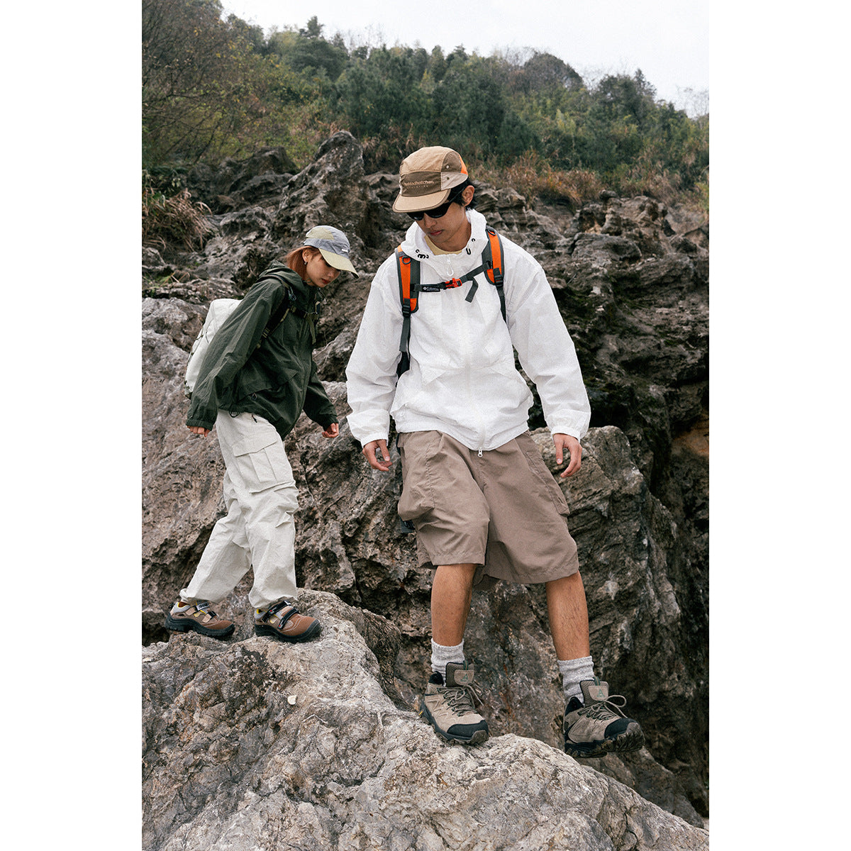 Two hikers on rocky terrain wearing outdoor layers; the man wears a white hooded jacket and taupe shorts.