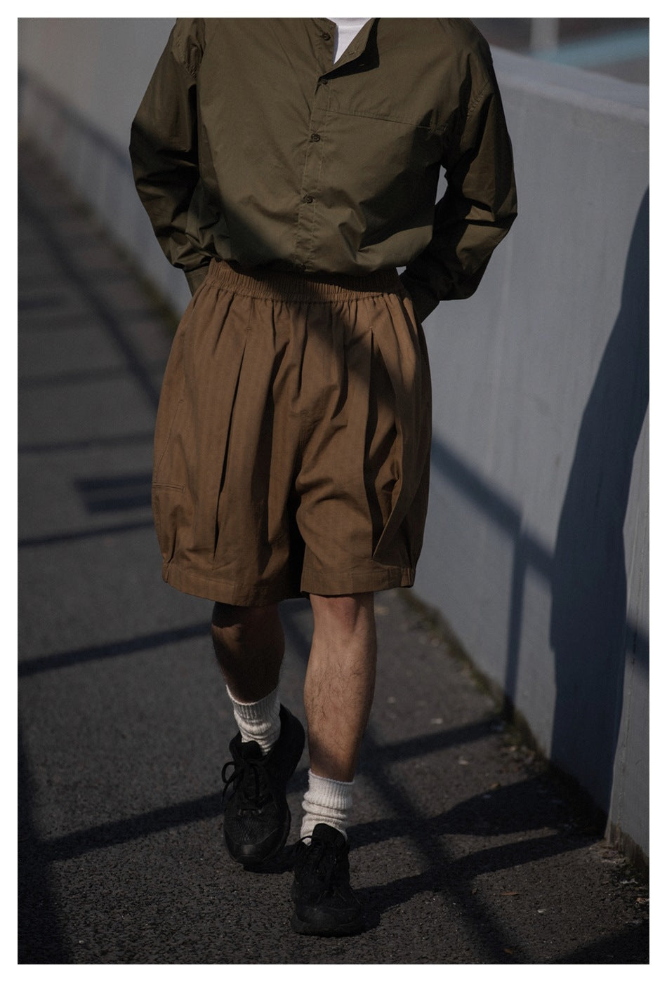 Cropped view of a model wearing brown loose shorts with an elastic waist and an olive button-up shirt.