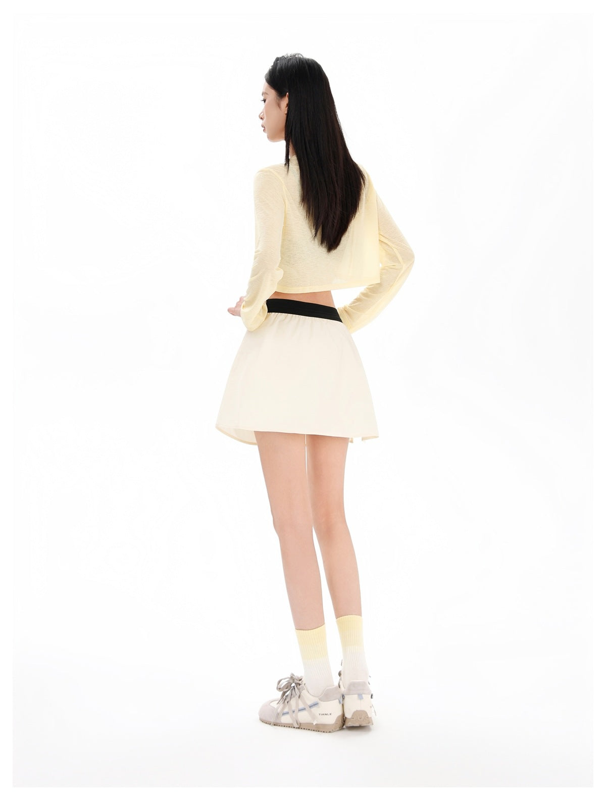 Rear view of a model wearing a pale yellow cropped long-sleeve top and cream flared skirt with a black waistband.