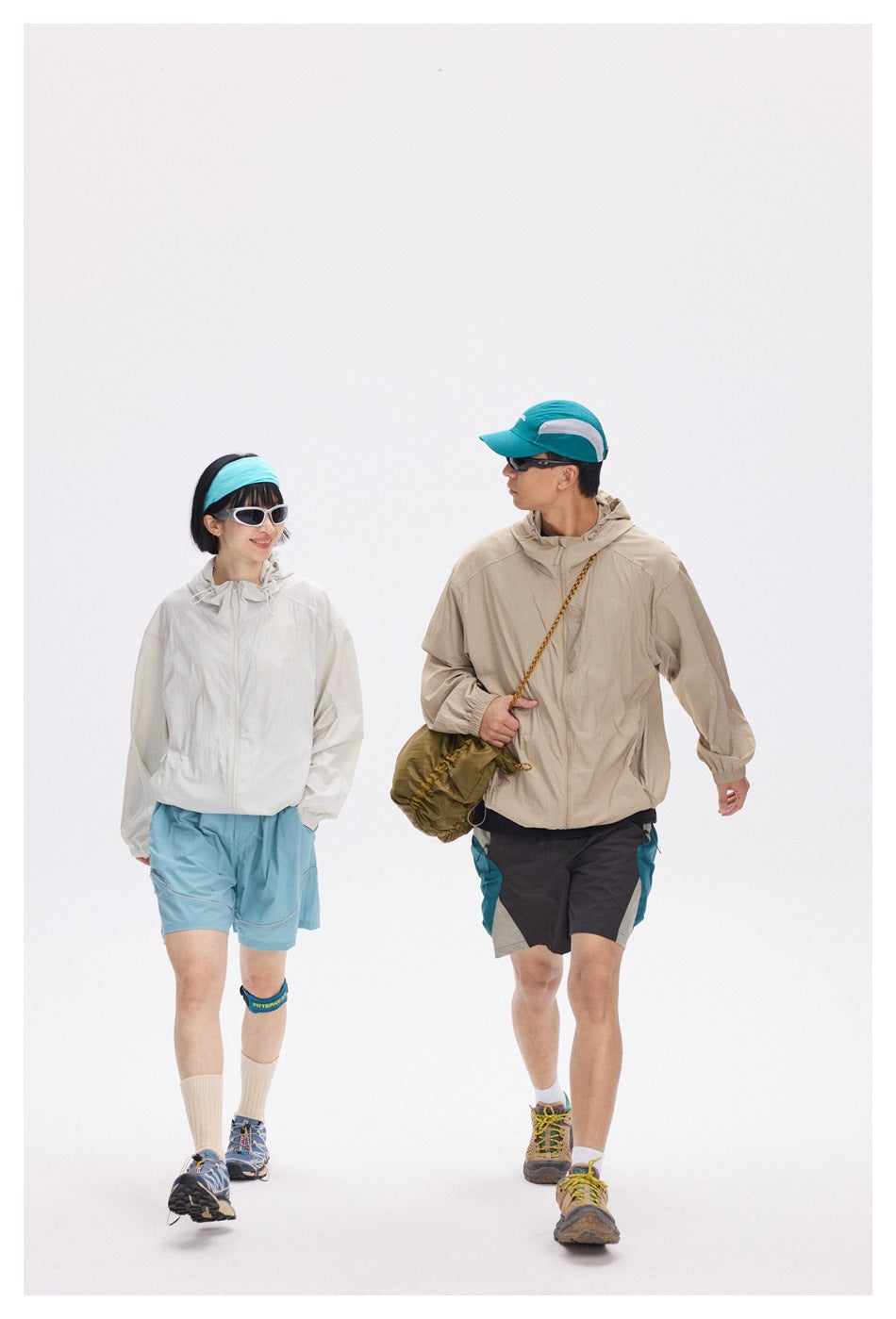 Two models walk in hiking outfits: an off-white jacket with blue shorts and a beige jacket with dark shorts.
