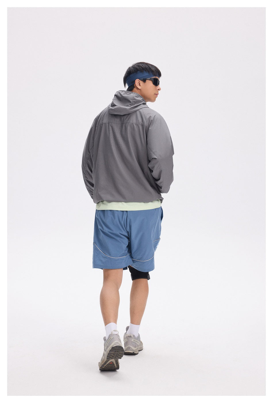 Rear view of a model wearing a charcoal gray hooded jacket with perforated back details and blue shorts.