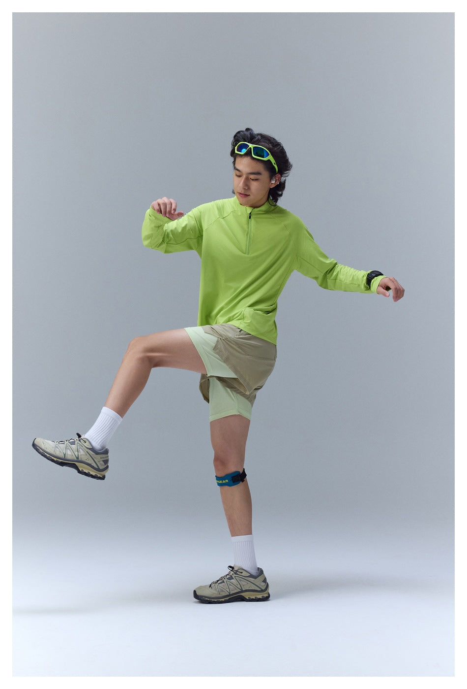 A man wears a bright lime quarter-zip top and layered beige and pale green shorts while posing.