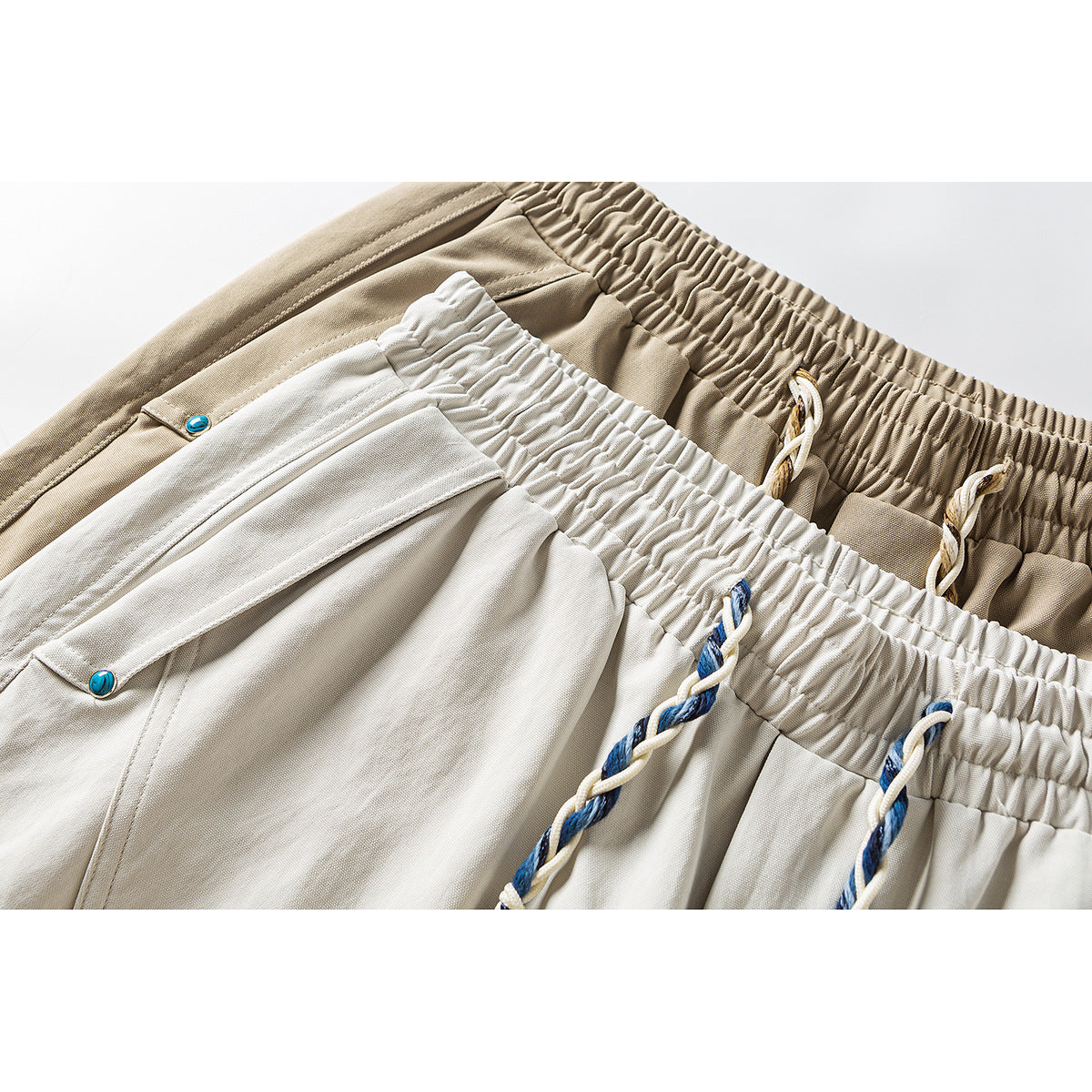 Close-up of light gray and taupe trousers showing elastic waistbands, braided drawstrings, and turquoise pocket rivets.