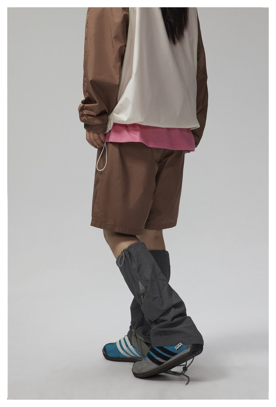Rear view of a person wearing a white jacket with brown sleeves, brown shorts, and dark gaiters over blue shoes.