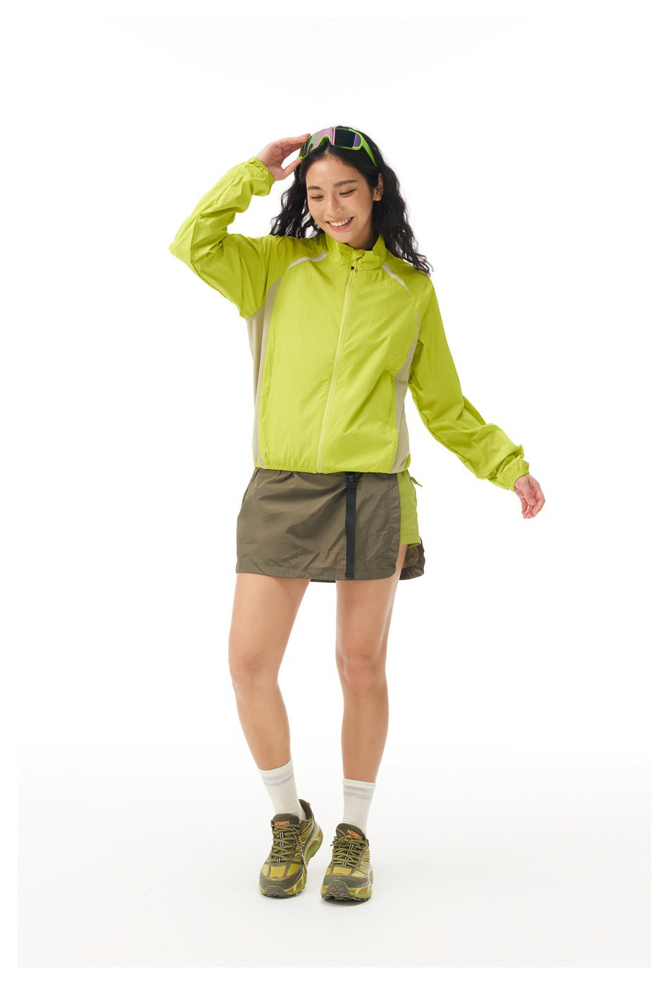 Woman modeling a lime-green zip jacket with light side panels and an olive hiking skirt against a white background.