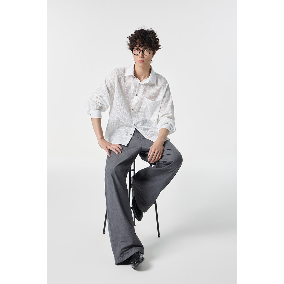 Seated model wearing a white textured button-up shirt with a chest pocket and gray wide-leg trousers.