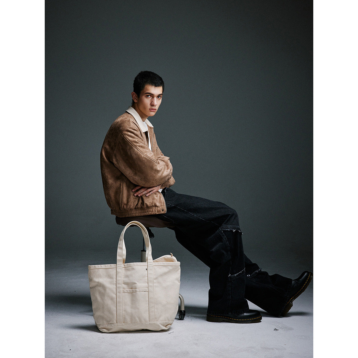 Seated man wearing a tan jacket with a cream collar and black jeans beside a large cream tote bag.
